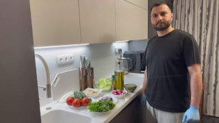 Человек на кухне у рабочей поверхности; на столе томаты, огурцы, пучки зелени, бутылки масла и подсушенный хлеб — подготовка к фаттушу.