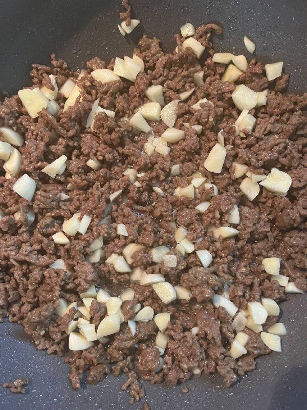 Обжаренный фарш с крупно нарезанным чесноком на сковороде — коричневая мясная масса и белые кусочки чеснока в процессе приготовления.