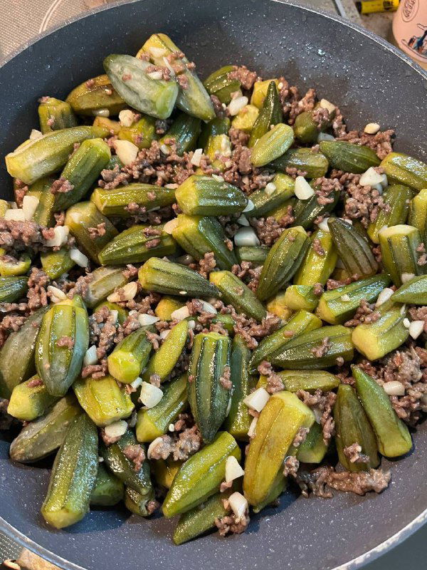 Сковорода с обжаренной бамией и фаршем — зелёные стручки перемешаны с кусочками мяса, видно сочетание ингредиентов перед тушением.