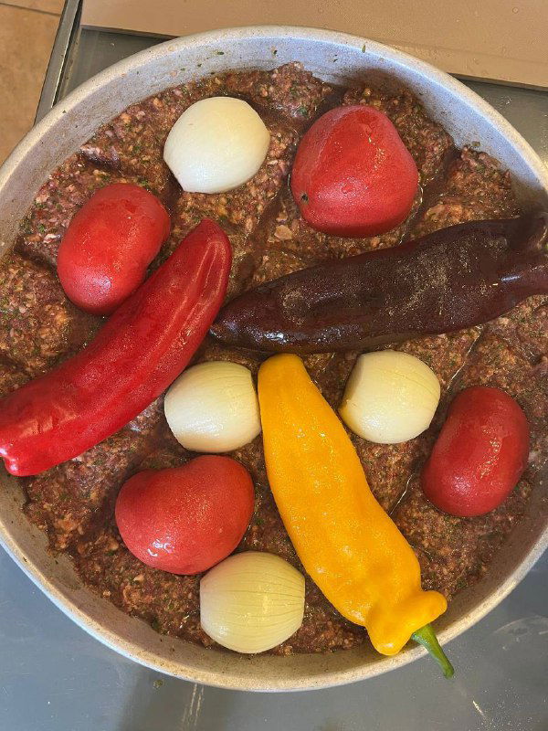 Собранный в противне фарш с поверхностью, на которой уложены целые помидоры, перцы рамиро и луковицы перед запеканием.