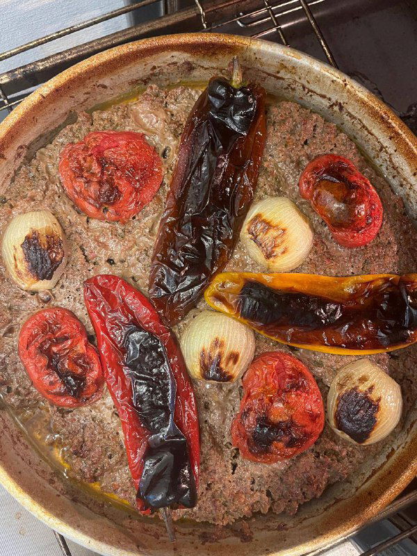 Противень после запекания: обугленные перцы, запечённые помидоры и лук на поверхности мясного пласта, видны карамелизация овощей.