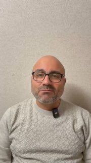 Портрет врача-гастроэнтеролога в очках и сером свитере на светлом нейтральном фоне; лицо смотрит в камеру, снимок статичный и информативный.