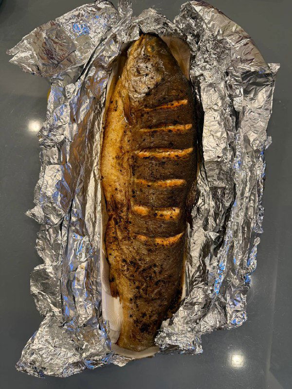 Запечённая целая рыбина в фольге на столе: видна золотистая корочка и надрезы, блюдо готово к подаче или на вынос после обработки и запекания