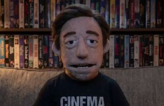 Пуговичный кукольный аватар мужчины в футболке с надписью CINEMA, тёплая кинотеатральная сцена и книжные полки на фоне.