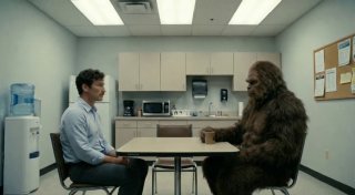 Офисная сцена переговоров: человек и большой гуманоид (сити‑креатура) за столом в комнате допросов — комедийный стейтмент в кадре.