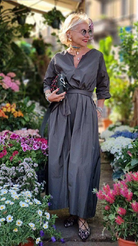 Женщина в тёмно‑сером платье‑халате среди цветов; струящаяся фактура и пояс создают элегантный праздничный силуэт для выпускного.