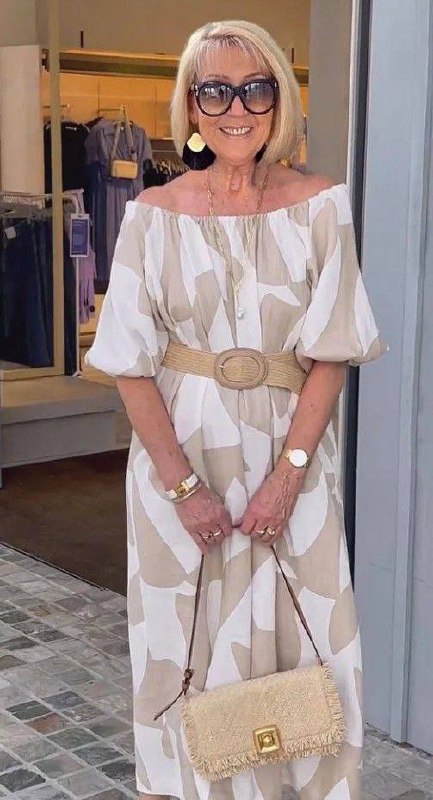 Женщина в пышном светлом платье с поясом и соломенной сумкой у входа в бутик; летний, женственный образ для праздничного вечера.