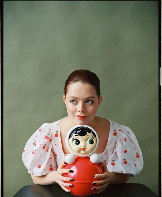 Портрет женщины в белой пышной блузе, она держит большую красную неваляшку у груди; студийный кадр с ретро‑настроением и фокусом на образе.