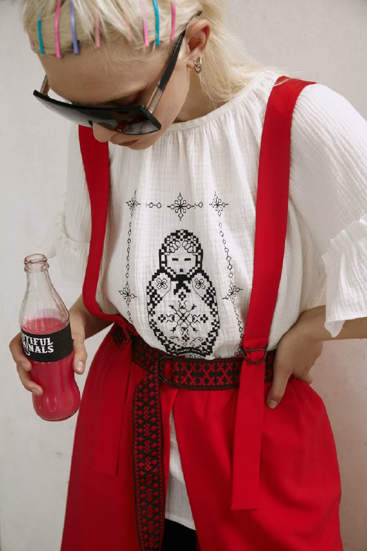 Уличный стиль: девушка в белой вышитой футболке с мотивом неваляшки и красных брейсов, образ с контрастным аксессуаром и бутылкой в руке.