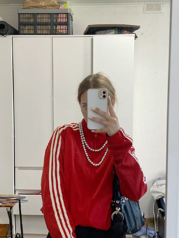 Зеркальное селфи: человек в красном спортивном костюме с двумя нитями жемчуга, телефон закрывает лицо, интерьерная обстановка, повседневный микс.