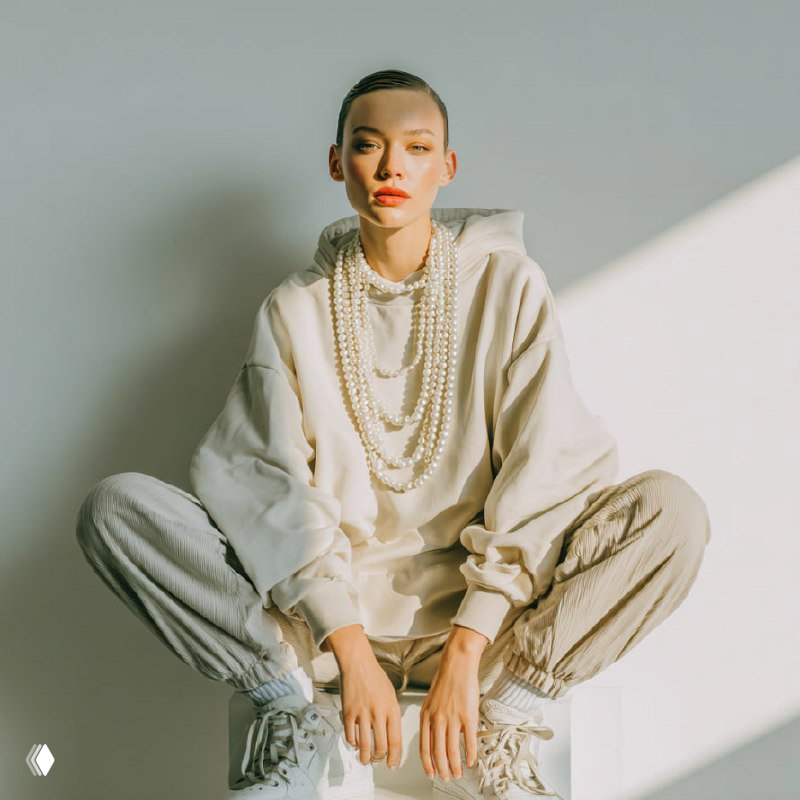 Студийный портрет модели в светлом худи и свободных брюках, многослойный жемчуг, поза с согнутыми коленями и мягкий боковой свет.
