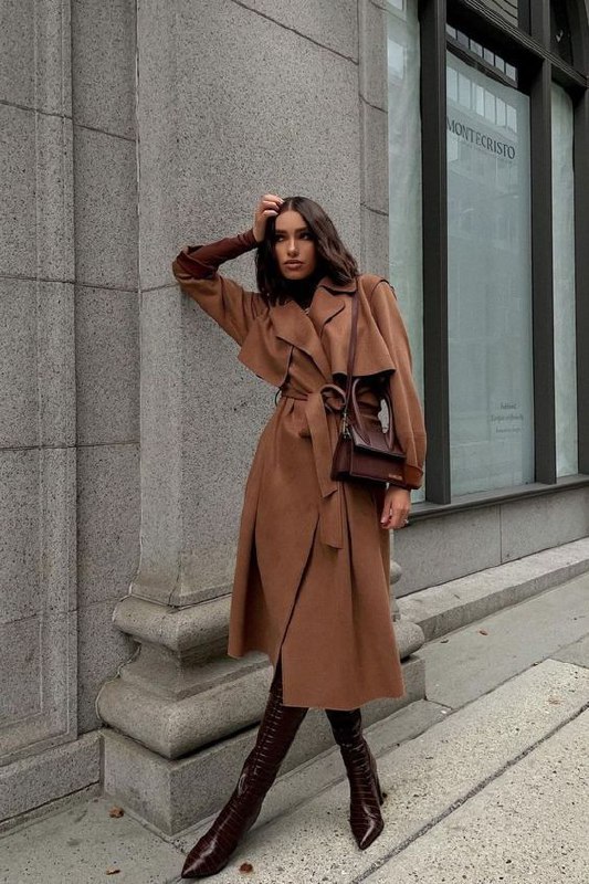Девушка в длинном коричневом тренче с поясом на фоне архитектуры — образ с акцентом на силуэт, движение и сдержанную элегантность.