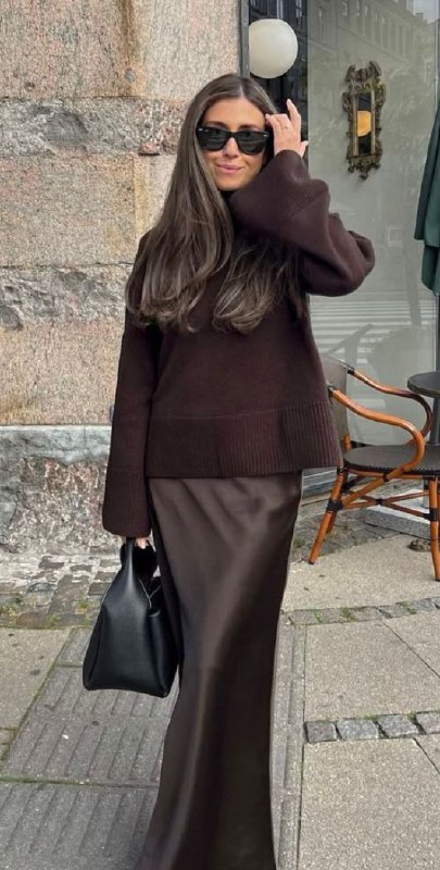 Женский образ в тёмно-коричневом свитере и длинной юбке, спокойная палитра и минималистичные аксессуары в урбанистическом окружении.