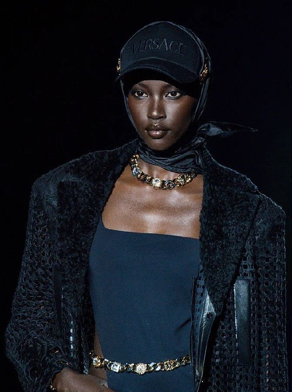 Модель на подиуме: тёмный лаконичный образ с платком на голове и заметным металлическим ожерельем, контраст кожи и аксессуаров подчёркнут светом