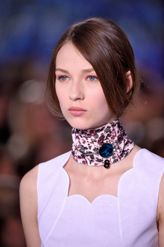 Модель на показе: платок или чокер вокруг шеи и крупный декоративный элемент у горла, светлый образ с чистым силуэтом и акцентом на шее