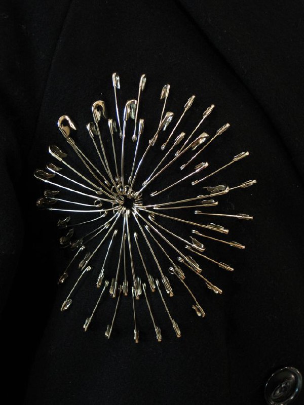 Брошь или декоративный элемент из множества маленьких серебристых булавок, собранных в круговую форму «фейерверка» на тёмном фоне, подчёркивающий текстуру.