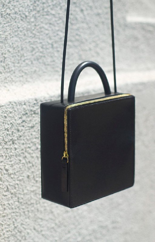 Чёрная квадратная сумка с короткой ручкой и золотой молнией на текстурном фоне; аксессуар-рамка, возвращающий ощущение устойчивости и упорядоченности.