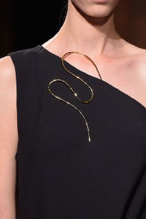 Золотое украшение в виде изгибающейся линии на чёрном платье, крупный план; плавная форма служит мягким, контактным акцентом в образе.