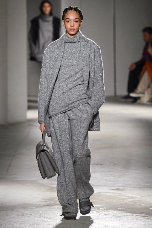 На подиуме модель в сером трикотажном костюме: свитер в однотон и широкие брюки, образ без лишней драматичности и сдержанный стиль.