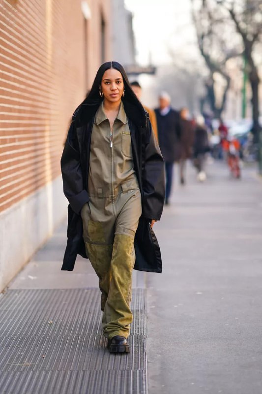 Модель в оливковом комбинезоне и тёмном пальто идёт по тротуару; пример лаконичного утилитарного стритстайла с монохромными тонами и грубой обувью.