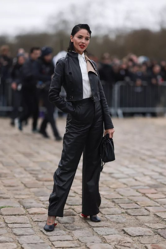 Девушка в чёрной кожаной куртке и кожаных брюках идёт по брусчатке перед толпой; минималистичный монохромный образ с акцентом на силуэт.