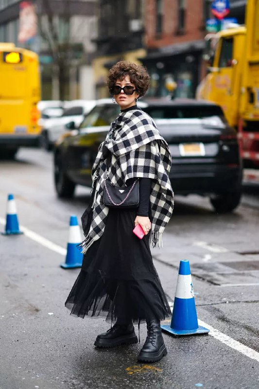 Женщина в длинной юбке с фатином и крупной клетчатой накидкой идёт по городу; эклектичное сочетание фактур и пропорций в уличном стиле.