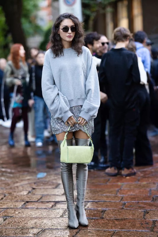 Женщина в сером объёмном свитере, серебристых сапогах и нежной сумкой стоит на городском фоне; контраст мужских форм и женских деталей в уличной моде.