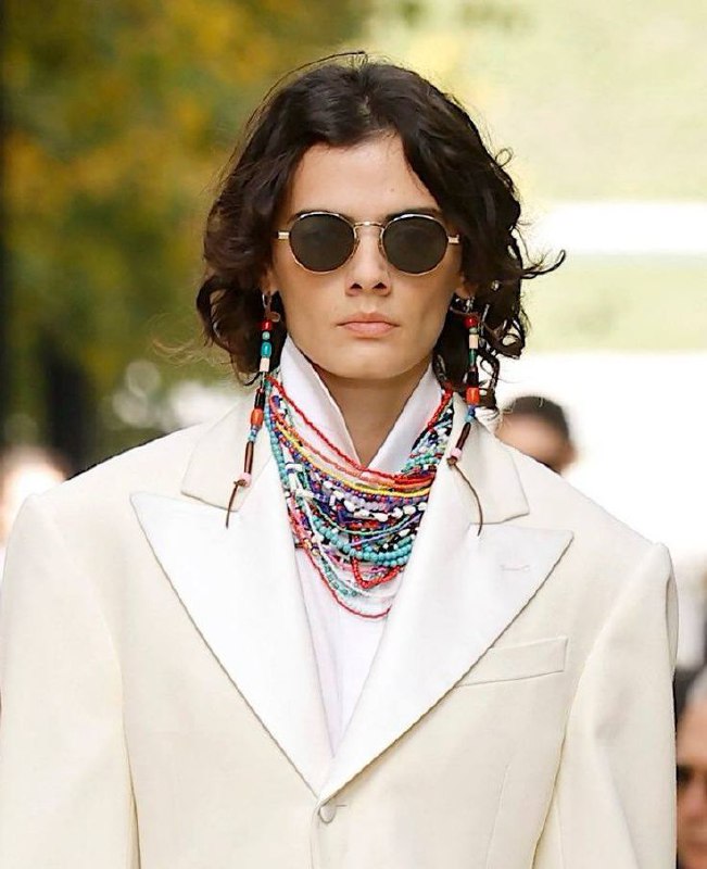 Модель в очках и большим количеством разноцветных ожерелий, комбинирование мужского пиджака и многоярусных бус, пример смешения стилей и повторов в аксессуарах.