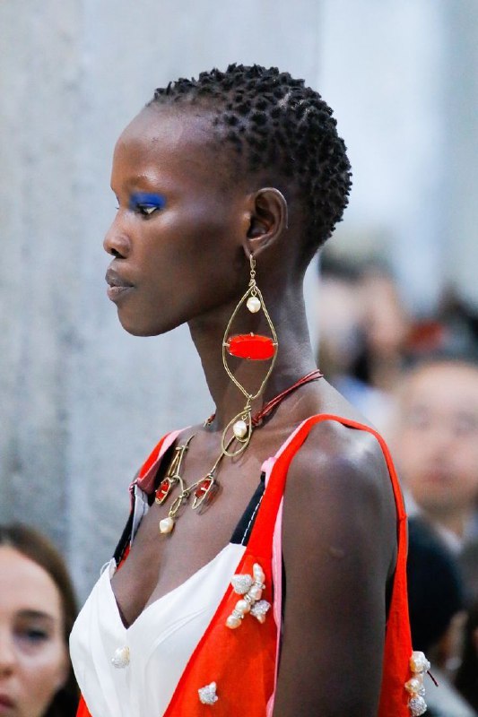 Темнокожая модель с массивными асимметричными украшениями и синими тенями, демонстрирующая контраст текста и форм в сложной аксессуарной композиции.