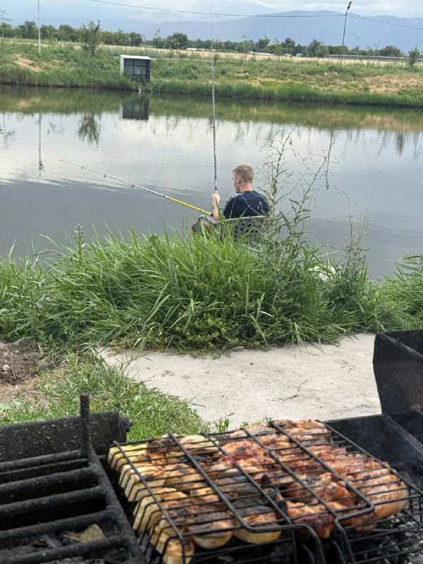 Гриль с множеством шампуров и приготовленным мясом на фоне озера и рыбака: бытовой кадр с обедом в дороге во время маршрута.