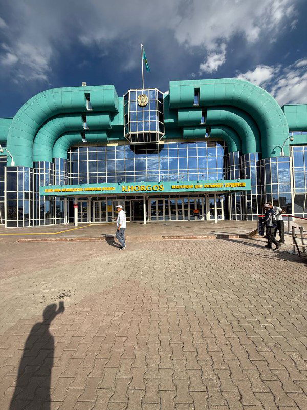 Яркое бирюзовое здание пограничного или логистического пункта с флагом на крыше и людьми у входа — остановка на границе Казахстана/Киргизии.