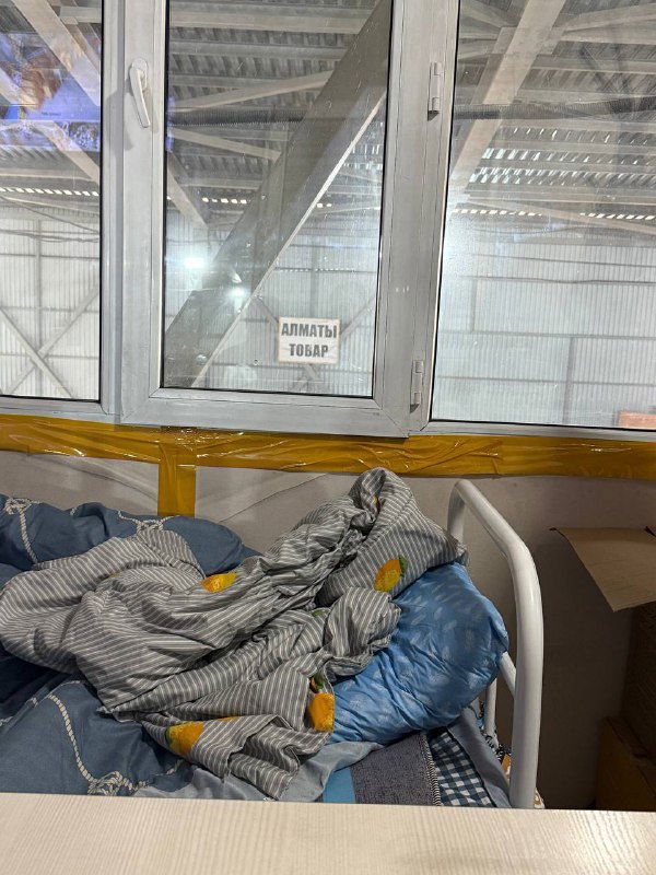 Внутренний кадр склада: окно с табличкой, рядом неубранное спальное место и одеяла — бытовой угол для водителей в логистическом хабе.