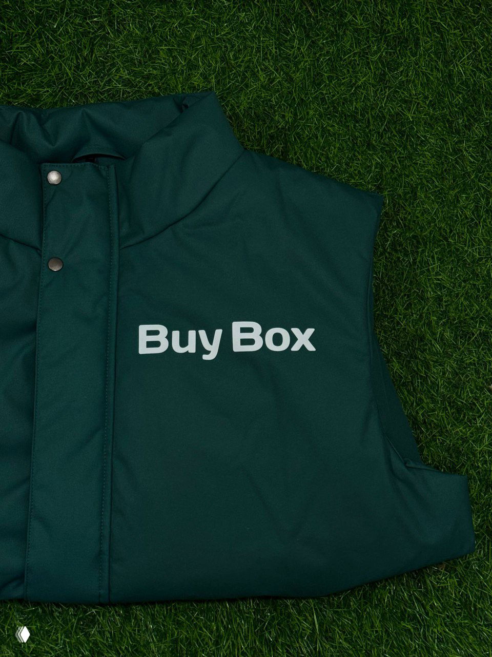 Тёмно-зелёная лёгкая жилетка, разложенная на траве; на груди крупная белая надпись «Buy Box», предмет мерча компании BuyBox.