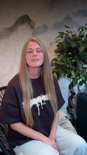 Превью: молодая женщина с длинными светлыми волосами сидит в кресле на фоне декоративной стены и большого комнатного растения, интерьерная портретная сцена.