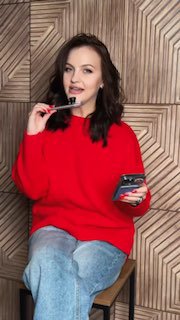 Женщина в красном свитере сидит на табурете перед деревянной панелью, держит смартфон в одной руке и аппликатор/щеточку в другой, улыбается.