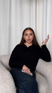 Кадр превью: женщина в тёмной одежде сидит на светлом кресле на фоне штор, жест рукой и нейтральное выражение — иллюстрация совета по оформлению карточки товара.