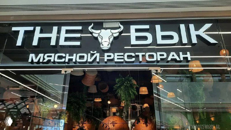 Вывеска и фасад мясного ресторана «The Бык!»: логотип с изображением быка, стеклянный вход и часть интерьера, видимые через витрину.