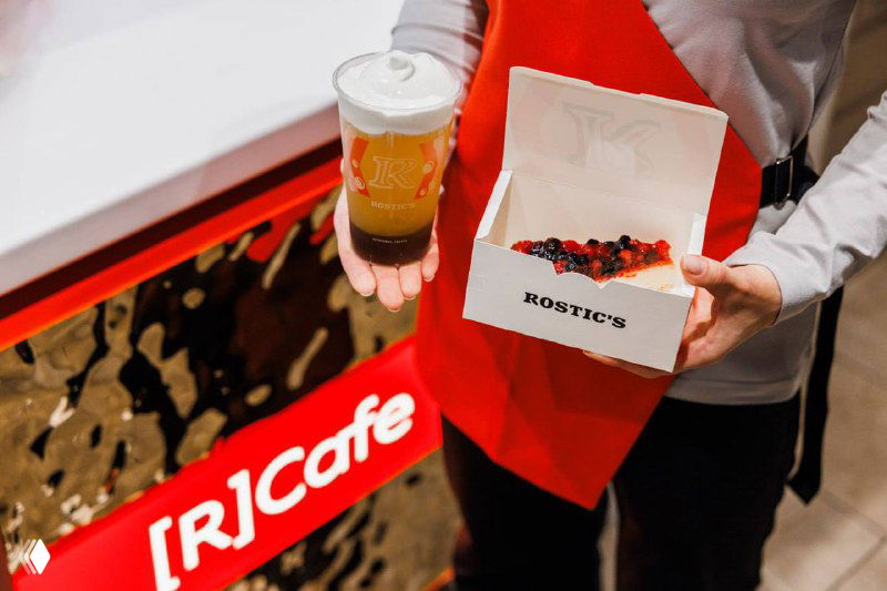 Сотрудник держит стакан с напитком и коробку с выпечкой Rostic’s: демонстрация ассортимента и брендированной упаковки в точке [R]Cafe в Москве
