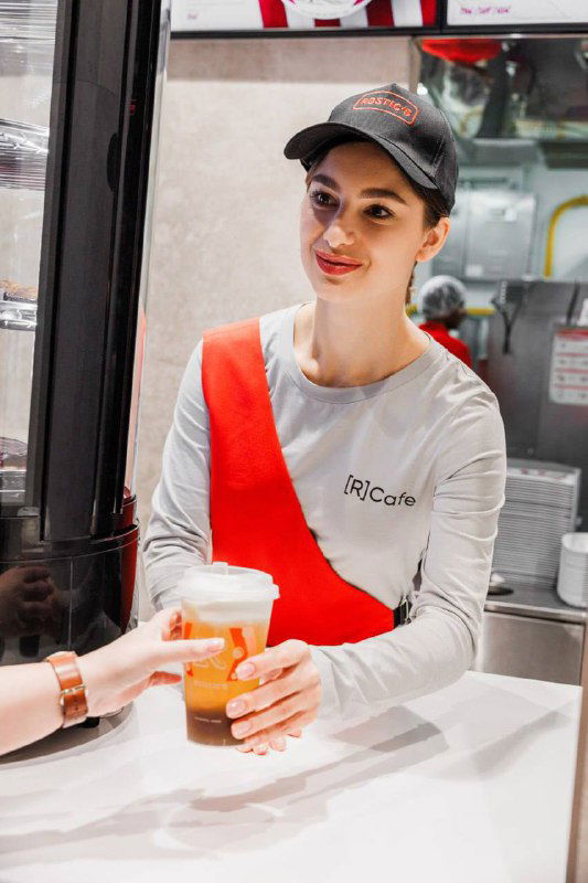 Бариста в форме [R]Cafe (фартук и кепка) подаёт напиток клиенту: кадр из московской пилотной точки сети Rostic’s, обслуживание и фирменная атрибутика