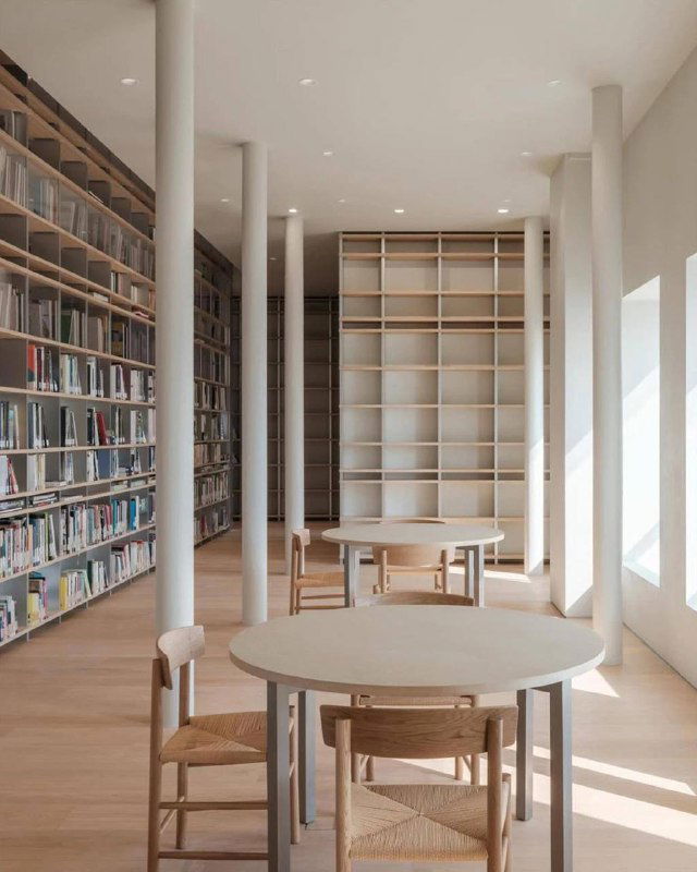 Интерьер читального зала с деревянными столами и стульями, большим количеством дневного света и аккуратной расстановкой мебели в спокойной палитре.