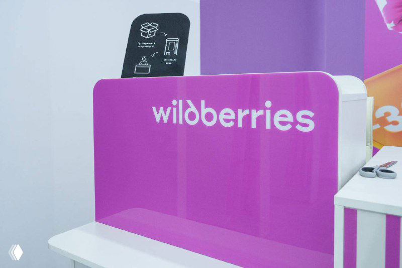 Деталь оформления стойки Wildberries с крупным белым логотипом на светло‑сиреневом фоне, демонстрация фирменной графики и материалов брендбука в интерьере.