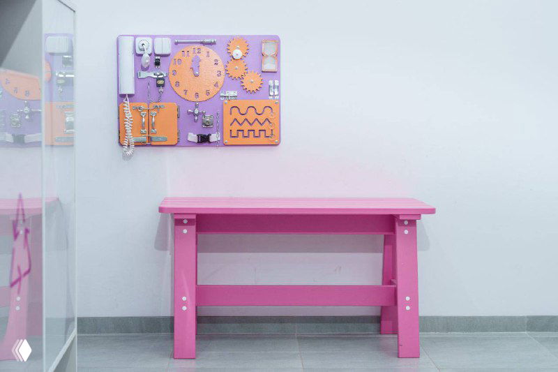 Детская зона внутри пункта выдачи: розовый столик и бизиборд на сиреневой панели, зона ожидания адаптирована для семей с детьми и короткого досуга.