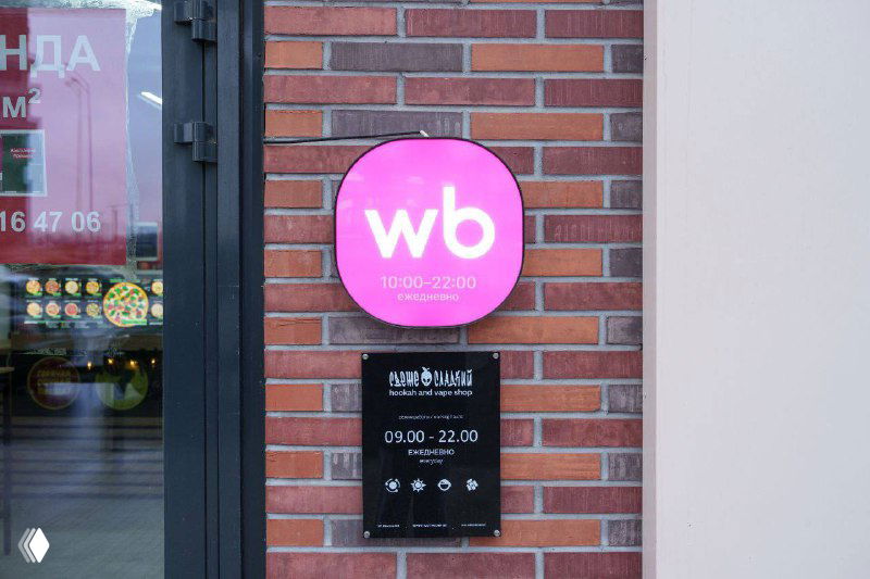 Крупный план круглого фирменного значка «wb» светло‑сиреневого тона и информационной таблички с режимом работы у входа в ПВЗ, элементы наружной навигации.