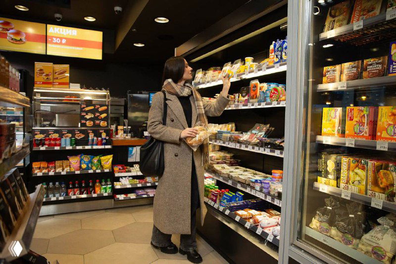 Посетитель выбирает товары с витрины в компактном магазине на АЗС: полки со снеком, упаковками и бакалеей на небольшом пространстве.