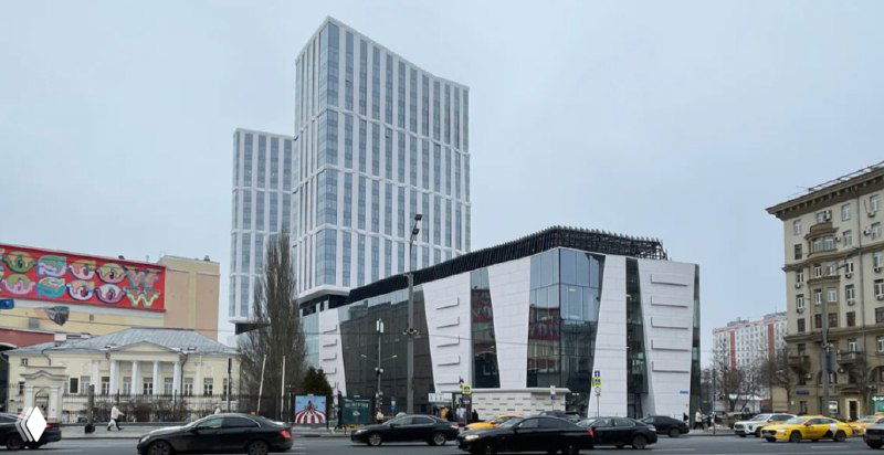 Фасад современного торгового центра на городской улице: входные группы, вывески и автомобильное движение на переднем плане.