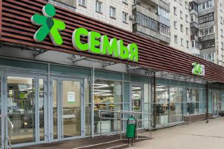 Вход в магазин с зеленой вывеской «Семья»: уличный фасад, стеклянные двери и элементы жилой застройки вокруг, фронтальный вид торговой точки.