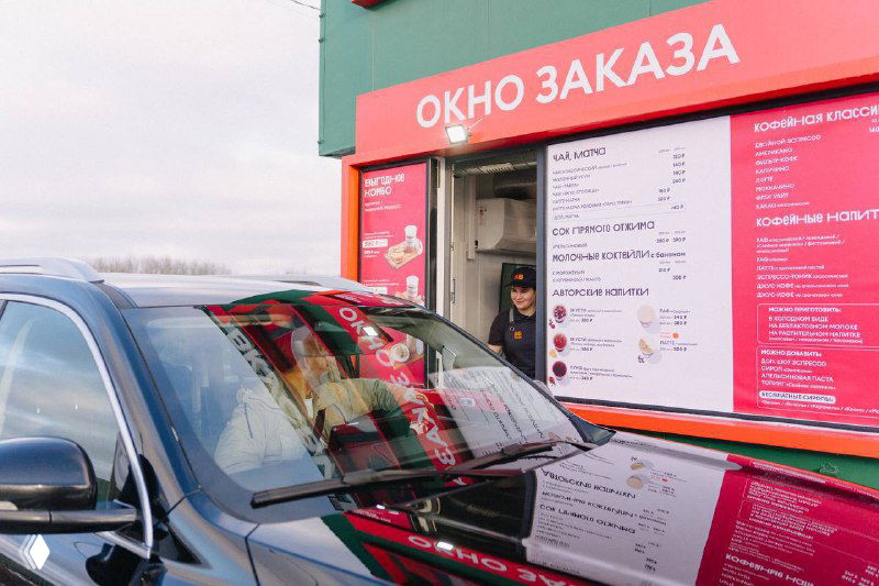 Окно заказа придорожного кафе: автомобиль у стойки, видна сотрудница в окне и меню на красочно оформленной панели рядом с входом.