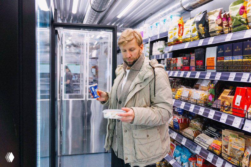 Внутренний вид минимаркета: мужчина с тарелкой у холодильника, стеллажи с товарами и упаковками, демонстрирующие ассортимент формата магазин+кафе.