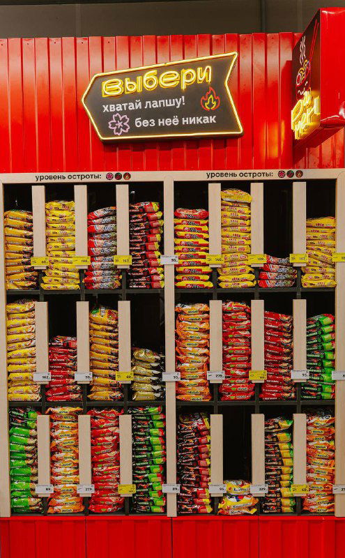 Стеллажи с ассортиментом упаковок лапши и топпингов в рамен‑баре «Магнит», красная вывеска с надписью «выбери лапшу» над витриной.