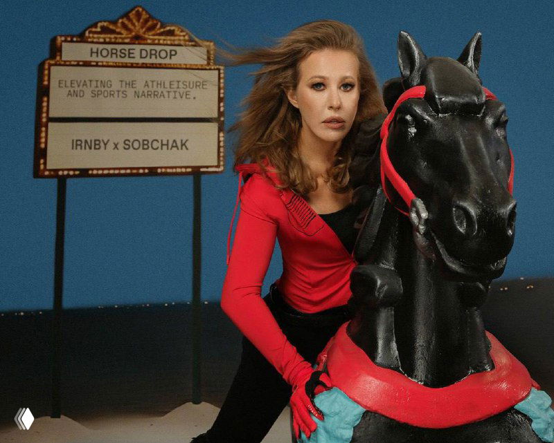 Кадр рекламной съёмки: женщина в красном образе рядом с чёрной статичной декорацией лошади в студии.